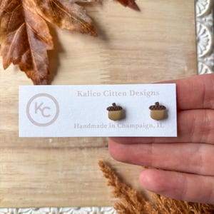 May include: A pair of small acorn-shaped stud earrings with dark brown caps and light tan bases. The earrings are attached to a white rectangular display card featuring the text Kalico Citten Designs and Handmade in Champaign, IL.