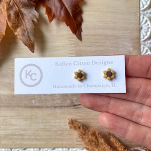 Puede incluir: Pendientes de botón de girasol sobre una tarjeta blanca. Los pendientes son amarillos y marrones, con un diseño floral detallado. La tarjeta dice "Kalico Citten Designs" y "Handmade in Champaign, IL."