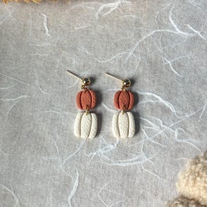 May include: A pair of drop earrings featuring a gold-tone post and two stacked pumpkin-shaped charms. The top pumpkin charm is a burnt orange color, while the bottom pumpkin charm is an off-white color. Both charms have a textured surface.