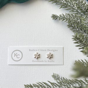 Könnte beinhalten: Silberne Schneeflocken-Ohrstecker auf einer weißen Karte mit dem Text "Kalico Citten Designs" und "Handmade by Kaylie". Die Ohrringe liegen auf einer strukturierten weißen Oberfläche, mit grünen Zweigen und einem grünen Stoff im Hintergrund.