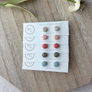 Pode incluir: Uma coleção de brincos de pressão em várias cores, incluindo bege, rosa, vermelho, verde-oliva e azul. Cada par é exibido em um pequeno cartão branco com o texto "Kalico Citten Designs" e "Handmade by Kaylie".