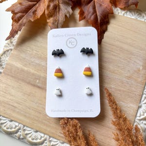 May include: A set of Halloween-themed stud earrings on a white card. The earrings include black bats, candy corn, and white ghosts. The card reads "Kalico Citten Designs" and "Handmade in Champaign, IL".