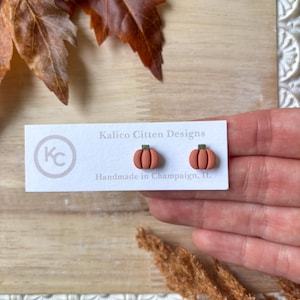 May include: A pair of terracotta-colored pumpkin stud earrings with green stems. The earrings are displayed on a white card with the text "Kalico Citten Designs Handmade in Champaign, IL". The background includes fall foliage.