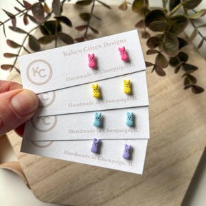 Op de afbeelding: Vier paar handgemaakte oorbel studs met pastelkleurige konijnenvormen. De oorbellen worden getoond op een witte kaart met de tekst "Kalico Citten Designs" en "Handmade in Champaign, IL".