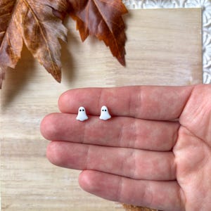 May include: A pair of small, white ghost-shaped stud earrings with black dot eyes. The earrings are displayed on a light wooden surface, with autumn leaves in the background. The earrings are a Halloween-themed accessory.