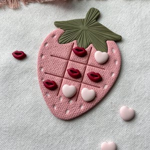 Può includere: Un gioco Tic-Tac-Toe a forma di fragola rosa con una parte superiore a forma di foglia verde. Il tabellone ha una griglia con pezzi di gioco a forma di labbra rosse e cuori rosa. Ulteriori pezzi a forma di labbra e cuori sono sparsi intorno al tabellone.