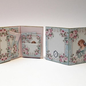 May include: Two miniature, open, decorative books with floral and portrait designs. The books feature a light blue background with pink roses and a gold trim. One book is open, revealing interior pages. The other book has a portrait of a woman.