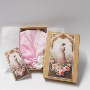 Peut inclure: Un article en tissu rose clair avec une bordure en dentelle blanche niché dans une petite boîte en carton. Le couvercle de la boîte et une carte rectangulaire séparée présentent une illustration d'une personne en robe longue parmi des roses.