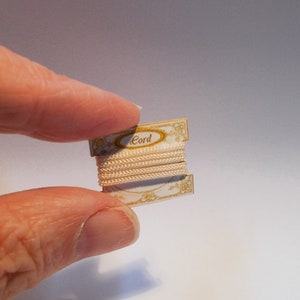 Könnte beinhalten: Eine kleine Spule weißen Kordels mit goldenen Verzierungen, auf der Verpackung steht "Cord".