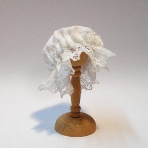 Op de afbeelding: Een witte kanten muts, waarschijnlijk voor een baby of pop, gepresenteerd op een kleine houten standaard. De muts heeft ingewikkelde kanten details rondom en is gemaakt van zacht, wit materiaal. De standaard is gemaakt van lichtgekleurd hout.