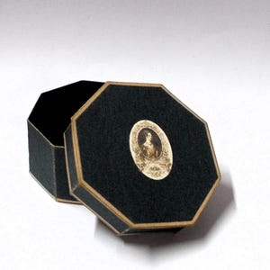 May include: An open, black, octagonal box with a gold trim. The lid features a small, oval portrait of a woman. The box is set against a white background. The box is likely used for jewelry or small keepsakes.