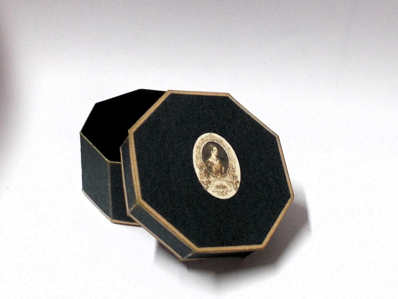 May include: An open, black, octagonal box with a gold trim. The lid features a small, oval portrait of a woman. The box is set against a white background. The box is likely used for jewelry or small keepsakes.