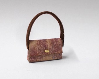 May include: A miniature handbag with a textured, reddish-brown exterior and a fuzzy, dark brown handle. The bag features a gold-colored clasp and a rectangular shape, suggesting a vintage or doll-sized accessory.