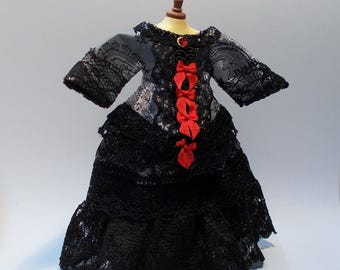 May include: A miniature black lace dress with long sleeves and a layered skirt. The bodice features a silver and black patterned fabric with red bows down the front and a red jewel at the neckline. The dress is displayed on a miniature mannequin.