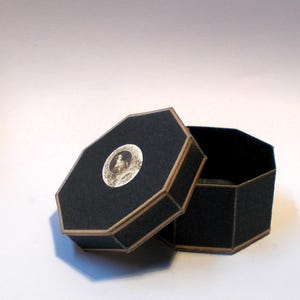 May include: An open, black, octagonal box with a hinged lid. The lid features a circular image of a person and a gold-colored trim. The box is on a white surface. The interior of the box is black.