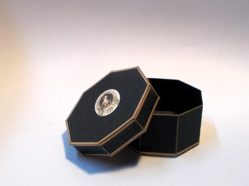 May include: An open, black, octagonal box with a hinged lid. The lid features a circular image of a person and a gold-colored trim. The box is on a white surface. The interior of the box is black.