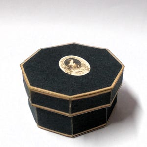 May include: A black, octagonal box with a gold trim. The lid features a small, oval-shaped image. The box is made of a dark, felt-like material. The box is likely used for jewelry or small keepsakes.