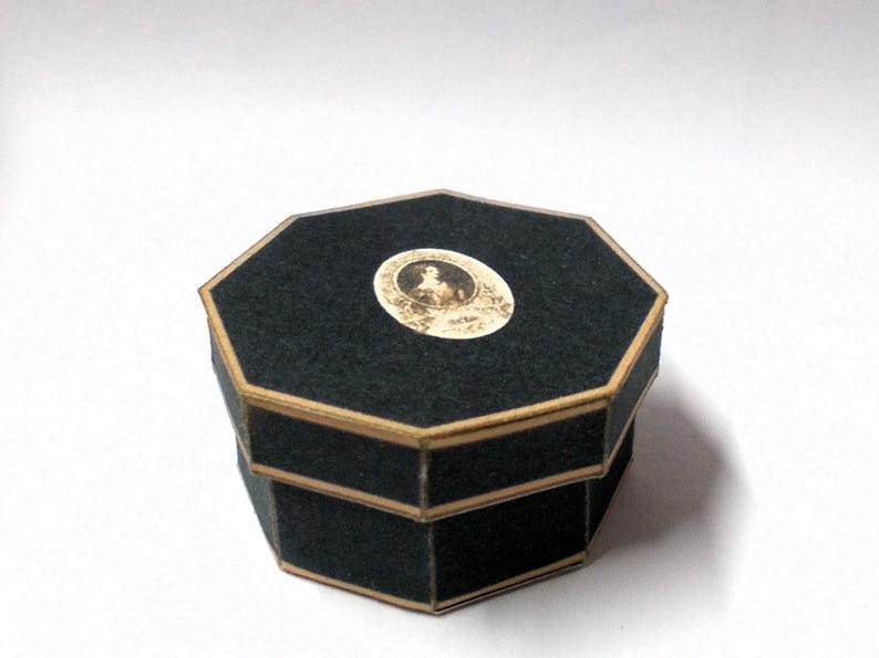 May include: A black, octagonal box with a gold trim. The lid features a small, oval-shaped image. The box is made of a dark, felt-like material. The box is likely used for jewelry or small keepsakes.