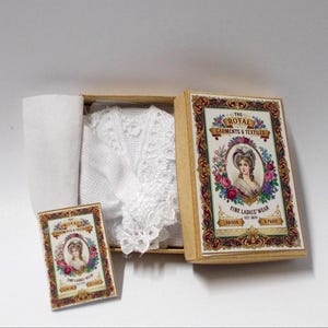 May include: A white crocheted garment inside a rectangular cardboard box with a vintage-style label featuring a portrait of a person, floral borders, and the text THE ROYAL GARMENTS & TEXTILES FINE LADIES' WEAR LONDON & PARIS EST 1870.