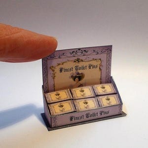 May include: A miniature display box of "Finest Toilet Pins" in a light purple hue. The box features six small, rectangular containers with a gold and cream design. The text "Finest Toilet Pins" is displayed on the front and top.