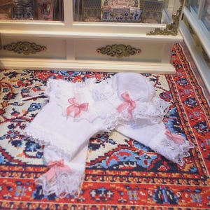 Op de afbeelding: Een witte poppenoutfit met kanten rand en roze strikken. De set bevat een top, broek en een muts. De outfit wordt gepresenteerd op een patroonkleed, wat wijst op een vintage of handgemaakte esthetiek. De details omvatten delicate kant en satijnen strikken.