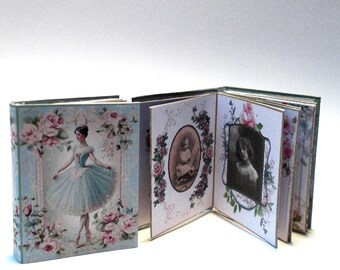 May include: Miniature handmade books with a vintage aesthetic. One book features a ballerina in a blue tutu surrounded by pink roses. The open book displays antique portraits framed by floral designs. The books are on a white surface.
