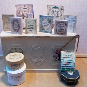 May include: A collection of miniature vintage-style books and decorative boxes. The books feature floral and botanical illustrations, with titles like "Embroidery Patterns" and "Hats & Bonnets." The boxes are round and hexagonal, with labels like "Maison de Paris."