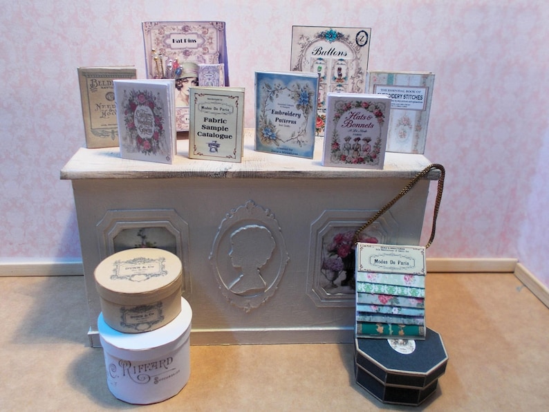 May include: A collection of miniature vintage-style books and decorative boxes. The books feature floral and botanical illustrations, with titles like "Embroidery Patterns" and "Hats & Bonnets." The boxes are round and hexagonal, with labels like "Maison de Paris."