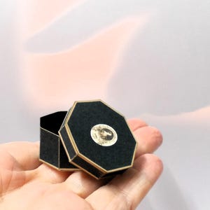 May include: A small, open black octagonal box with gold trim and a circular image on the lid. The box is held in a person's hand against a soft, blurred background.