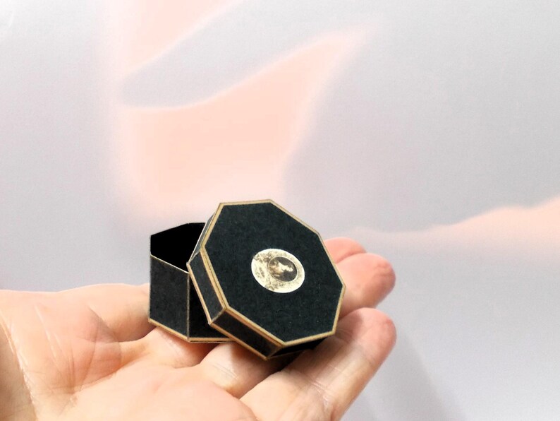 May include: A small, open black octagonal box with gold trim and a circular image on the lid. The box is held in a person's hand against a soft, blurred background.