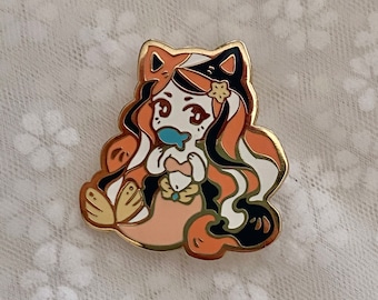May include: A gold-toned enamel pin featuring a cartoon mermaid with cat ears. The mermaid has orange, black, and white hair, a peach-colored top, and a blue fish. The pin has a gold border and a butterfly tail.