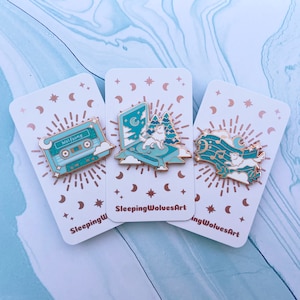 Könnte beinhalten: Drei Emaille-Pins mit einer Kassette, einem Wolf, der zum Mond heult, und einem schlafenden Wolf auf einer Musiknote. Die Pins befinden sich auf weißen Karten mit einem Muster aus goldenen Sternen und Halbmonden. Der Text "Sleeping Wolves Art" ist auf den Karten gedruckt.