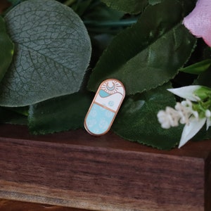Puede incluir: Un pin de esmalte azul claro y blanco con forma de píldora con un diseño de sol y luna. El pin tiene un fondo blanco con una ola azul y un diseño de sol y luna en la mitad superior. La mitad inferior del pin es azul claro con un patrón floral blanco.