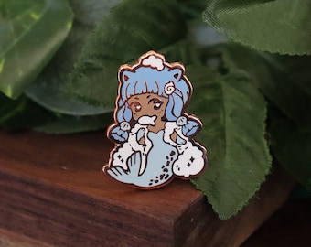 May include: A blue and white enamel pin featuring a cartoon mermaid with a cloud on her head. She is wearing a white fur coat and holding a fish in her mouth.