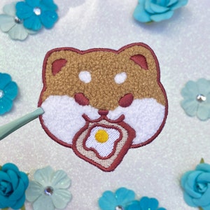 May include: An embroidered patch featuring a Shiba Inu dog, depicted in brown and white, enjoying a fried egg. The patch is crafted from soft, fluffy fabric and has a charming, cartoon-like design.