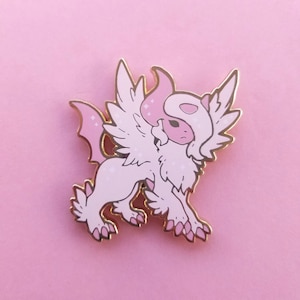 May include: A pink and gold enamel pin depicting a mythical creature with wings, a fluffy tail, and a pink and white head. The creature has a gold outline and sparkles.