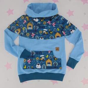 Könnte beinhalten: Ein hellblauer Kapuzenpullover mit dunkelblauer Passe und Fronttasche mit Bauernhof-Motiv. Das Muster zeigt Kühe, Schafe, Hühner, gelbe Traktoren, Scheunen und Sonnenblumen. Das Kleidungsstück hat einen Wasserfallkragen, lange Ärmel und Bündchen.