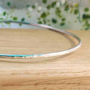 May include: A silver bangle bracelet with a hammered texture. The bracelet is thin and delicate, and it is shown on a wooden surface.