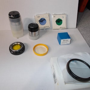 May include: An assortment of telescope accessories, including eyepieces, filters, and a blue Meade box. The items are arranged on a white surface. Two Tiffen filters are visible, along with a black and silver eyepiece.