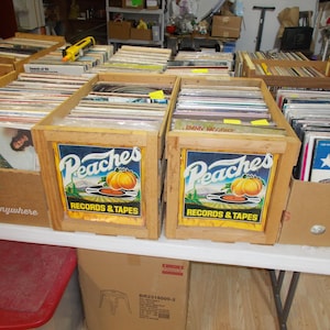 May include: Wooden crates filled with vinyl records. The crates have a "Peaches Records & Tapes" logo featuring a pumpkin. The records are in various sleeves, some with visible album art. The crates are on a white table.