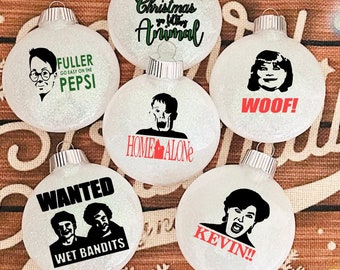 Può includere: Una collezione di sei palline di Natale trasparenti e glitterate. Ogni pallina presenta una grafica e un testo in nero e rosso tratti dal film "Mamma, ho perso l'aereo". Le frasi includono "Fuller, Go Easy on the Pepsi", "Merry Christmas ya Filthy Animal", "Woof!", "Home Alone", "Wanted Wet Bandits" e "Kevin!!".