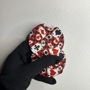 Könnte beinhalten: Zwei Auto-Untersetzer mit Spielkarten-Design. Die Untersetzer haben ein rot-schwarzes Karomuster mit Spielkartenfarben, darunter Herzen, Rauten, Kreuz und Pik. Die Untersetzer werden von einer Person mit einem schwarzen Handschuh gehalten.