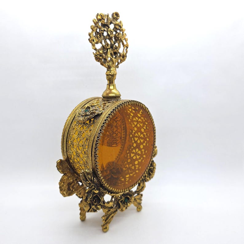 May include: A vintage gold-toned perfume bottle with a decorative floral stand. The bottle has a round amber-colored glass center with a filigree design. The top features a tree-like ornament.
