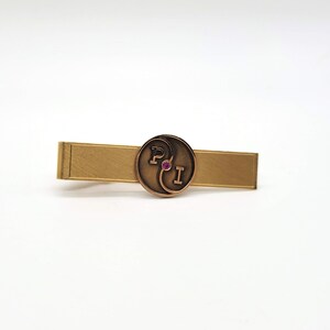 May include: A gold tie bar with a circular design featuring the letters "P" and "I" in a stylized font. A small pink stone is centered in the design.