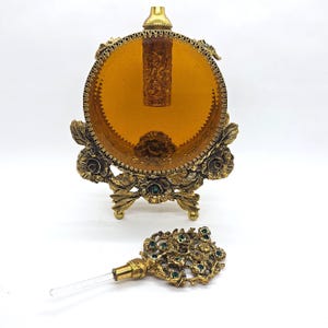 May include: An antique perfume bottle with a decorative gold-tone stand and a matching stopper. The stand features floral and leaf designs with green accents. The bottle itself is amber-colored and cylindrical, with intricate carvings. The stopper is also gold-toned with a floral design.