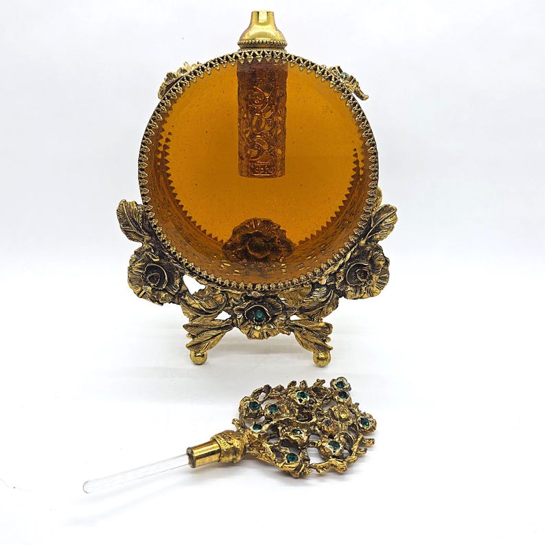 May include: An antique perfume bottle with a decorative gold-tone stand and a matching stopper. The stand features floral and leaf designs with green accents. The bottle itself is amber-colored and cylindrical, with intricate carvings. The stopper is also gold-toned with a floral design.
