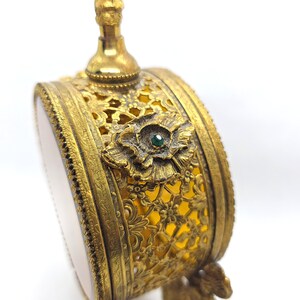 May include: Ornate antique perfume bottle with a gold-tone filigree design. The bottle features a floral embellishment with a small green gemstone at its center. The bottle has a round shape and a decorative top.