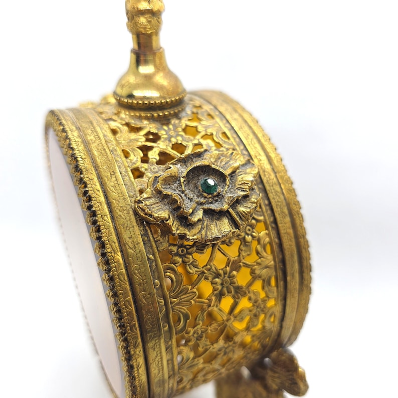 May include: Ornate antique perfume bottle with a gold-tone filigree design. The bottle features a floral embellishment with a small green gemstone at its center. The bottle has a round shape and a decorative top.