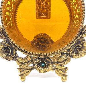 May include: Ornate, vintage vanity tray with a golden hue. The tray features a round, amber-colored glass insert surrounded by a decorative, filigree metal frame. The base is supported by floral and leaf-shaped legs, with a small green jewel accent.