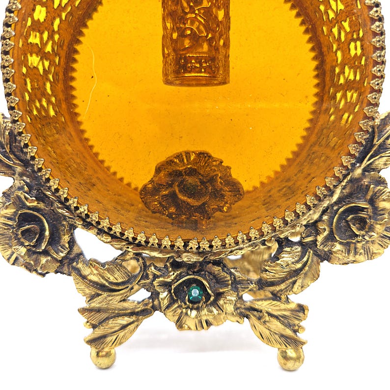 May include: Ornate, vintage vanity tray with a golden hue. The tray features a round, amber-colored glass insert surrounded by a decorative, filigree metal frame. The base is supported by floral and leaf-shaped legs, with a small green jewel accent.
